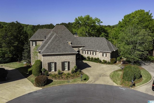 french country home with a view of trees, brick siding, curved driveway, and roof with shingles