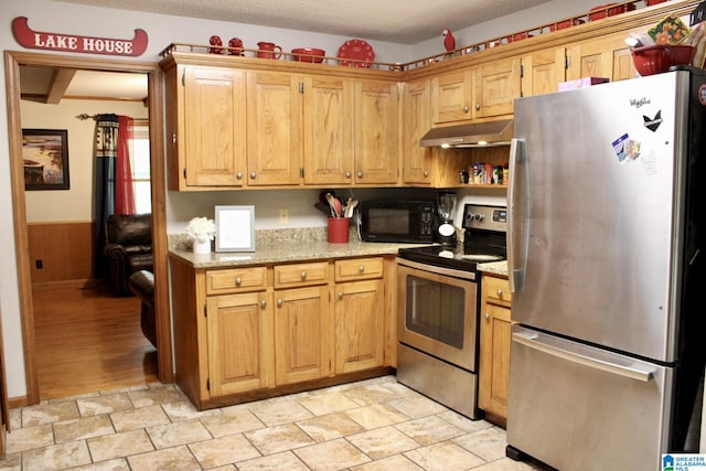 kitchen featuring under cabinet range hood, appliances with stainless steel finishes, stone finish flooring, a wainscoted wall, and a textured ceiling