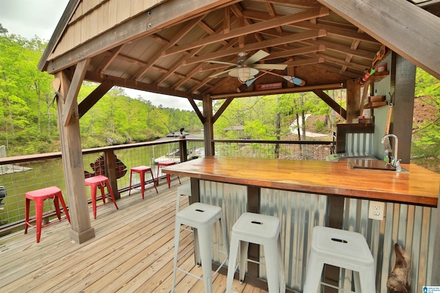 wooden terrace featuring a gazebo, outdoor dry bar, a sink, a ceiling fan, and a view of trees
