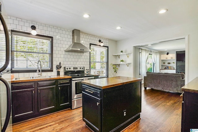 kitchen with butcher block countertops, wall chimney range hood, dark cabinetry, a sink, and appliances with stainless steel finishes