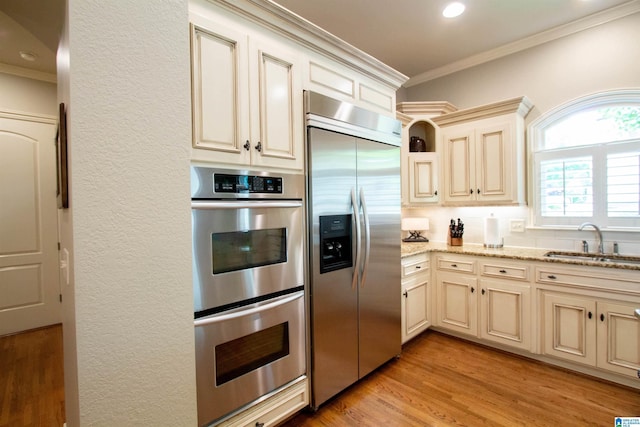 kitchen with appliances with stainless steel finishes, a sink, cream cabinetry, ornamental molding, and light stone counters