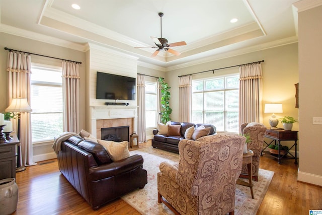 living room with a tray ceiling, light wood-style floors, ceiling fan, a tiled fireplace, and crown molding