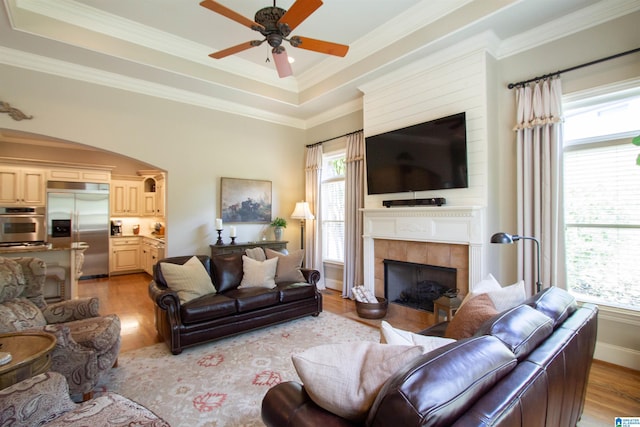living room featuring light wood-type flooring, a fireplace, ceiling fan, a raised ceiling, and ornamental molding