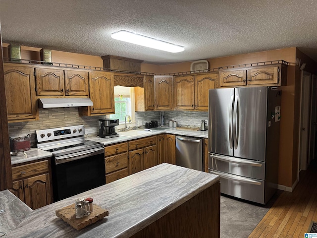 kitchen featuring stainless steel appliances, a textured ceiling, a sink, brown cabinets, and under cabinet range hood