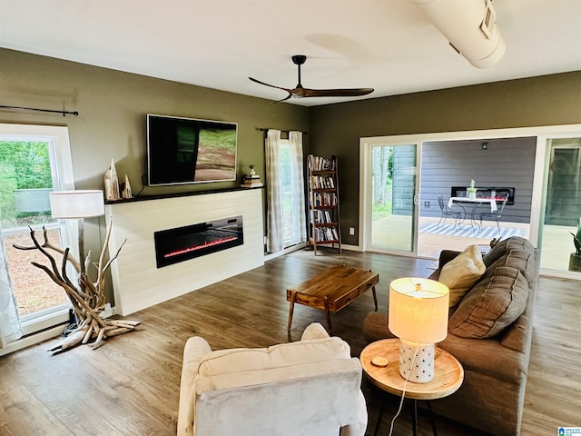 living area featuring a ceiling fan, a healthy amount of sunlight, wood finished floors, and a glass covered fireplace