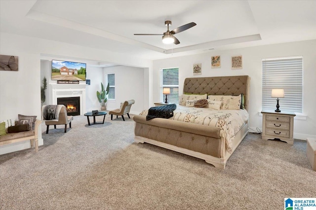 carpeted bedroom with baseboards, ceiling fan, a lit fireplace, and a tray ceiling