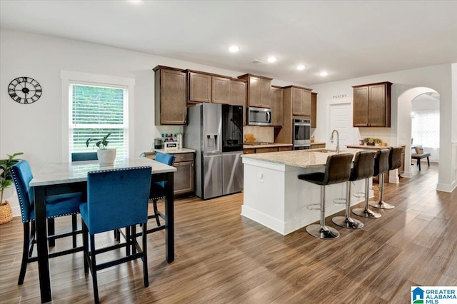 kitchen with arched walkways, a kitchen island with sink, wood finished floors, appliances with stainless steel finishes, and a sink