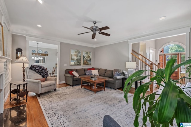 living room with a fireplace, ceiling fan with notable chandelier, stairs, wood finished floors, and ornamental molding