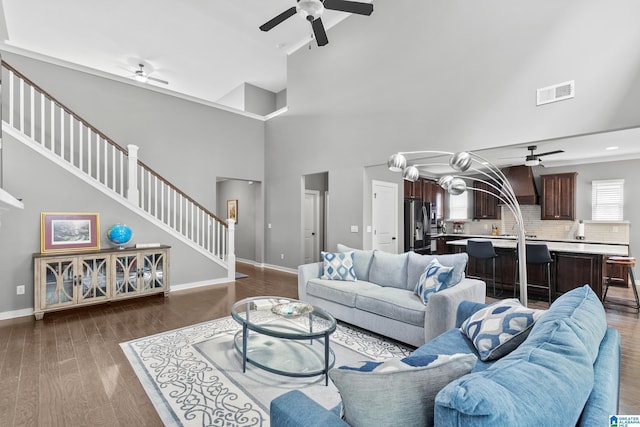 living area with a towering ceiling, a ceiling fan, dark wood-type flooring, visible vents, and stairway