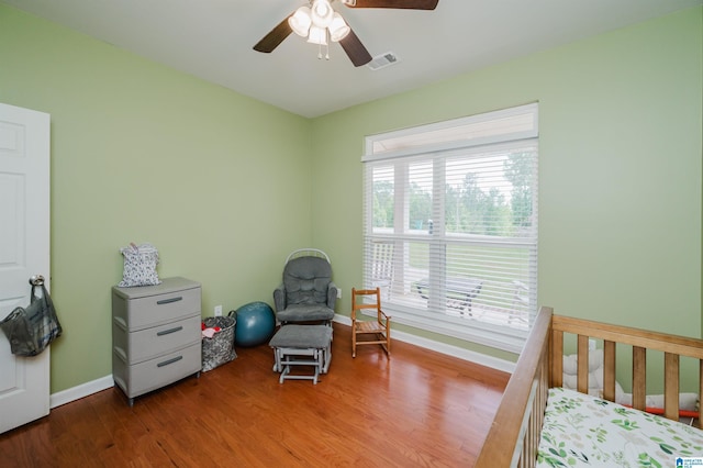 bedroom with ceiling fan, visible vents, a nursery area, wood finished floors, and baseboards