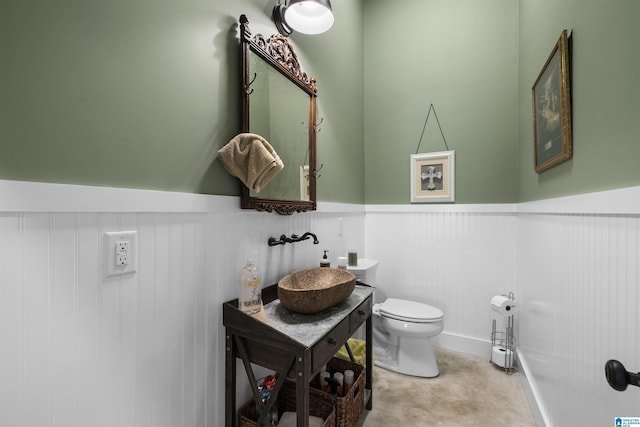 half bath featuring vanity, concrete flooring, a wainscoted wall, and toilet