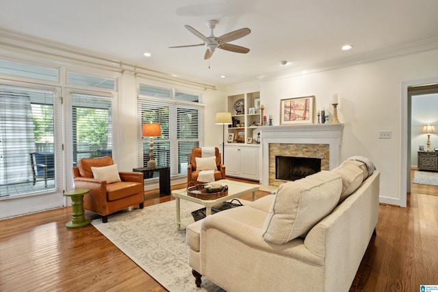 living room with crown molding, wood finished floors, a stone fireplace, recessed lighting, and a ceiling fan