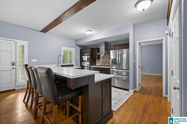 kitchen featuring dark brown cabinetry, a sink, light countertops, wall chimney exhaust hood, and stainless steel appliances
