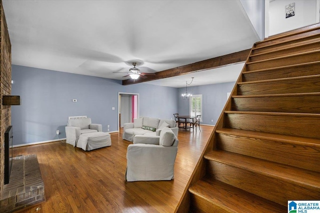 living room featuring a brick fireplace, wood finished floors, stairway, and ceiling fan with notable chandelier