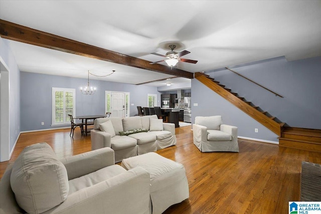 living area featuring ceiling fan with notable chandelier, baseboards, wood finished floors, beamed ceiling, and stairway