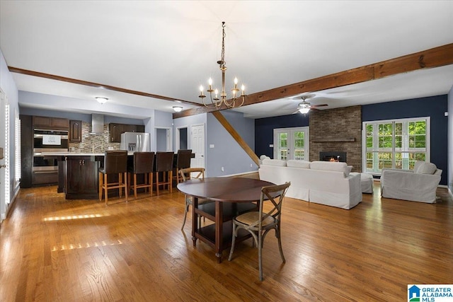 dining room with beam ceiling, wood finished floors, ceiling fan with notable chandelier, and a fireplace