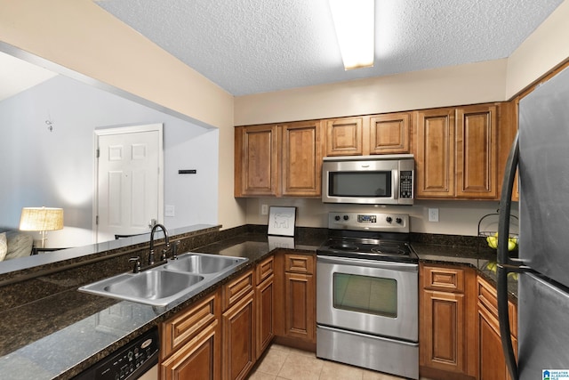 kitchen with brown cabinets, appliances with stainless steel finishes, a sink, a textured ceiling, and dark stone countertops