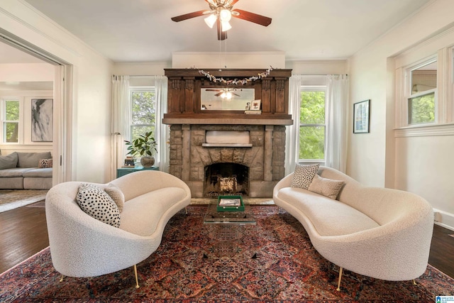 living area with plenty of natural light, wood finished floors, ceiling fan, a stone fireplace, and crown molding