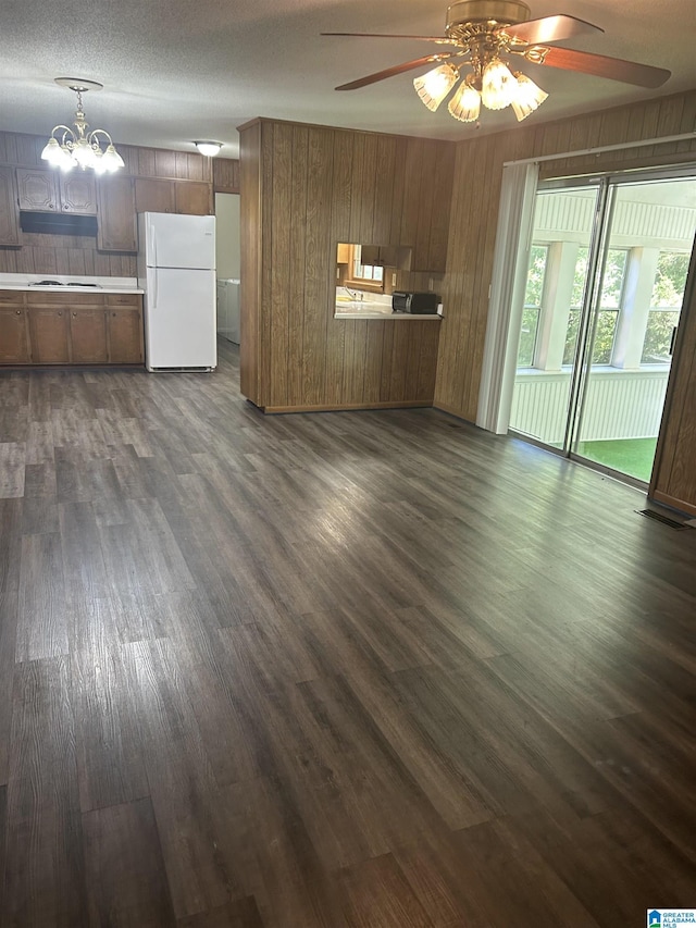unfurnished living room featuring wood walls, ceiling fan with notable chandelier, and dark wood-type flooring