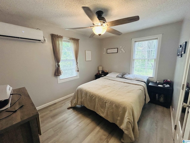 bedroom with a ceiling fan, a textured ceiling, a wall mounted AC, baseboards, and light wood-style floors