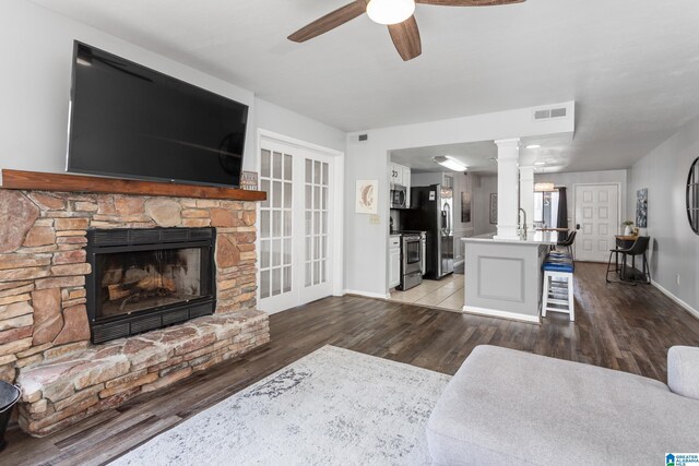 living room featuring wood finished floors, a ceiling fan, baseboards, a stone fireplace, and ornate columns