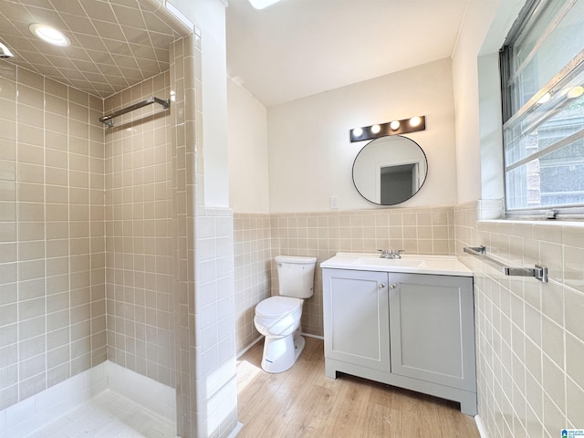 bathroom with tile walls, wood finished floors, toilet, tiled shower, and vanity