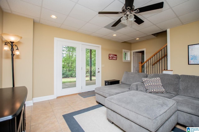 living area with a paneled ceiling, ceiling fan, light tile patterned flooring, stairway, and baseboards