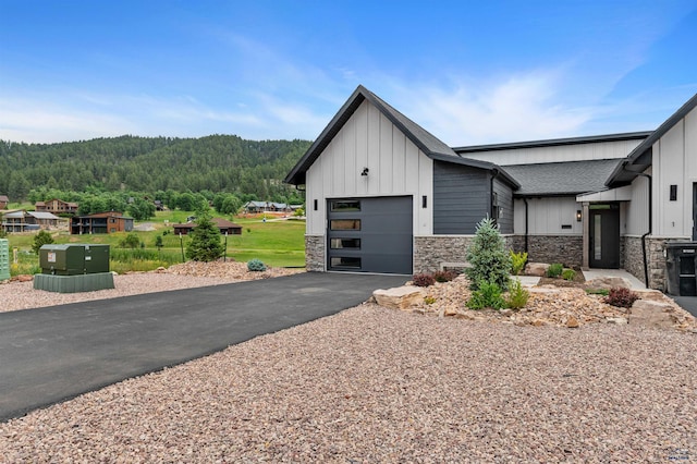 view of front of property featuring a wooded view, board and batten siding, aphalt driveway, stone siding, and an attached garage