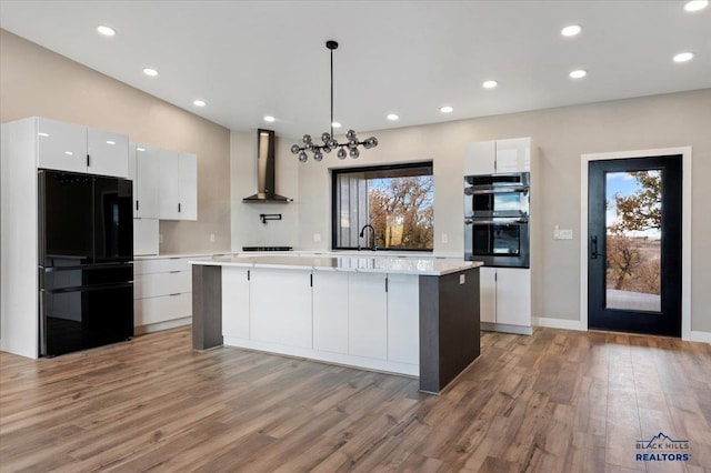 kitchen with wall chimney range hood, white cabinetry, black appliances, light wood-style flooring, and a kitchen island