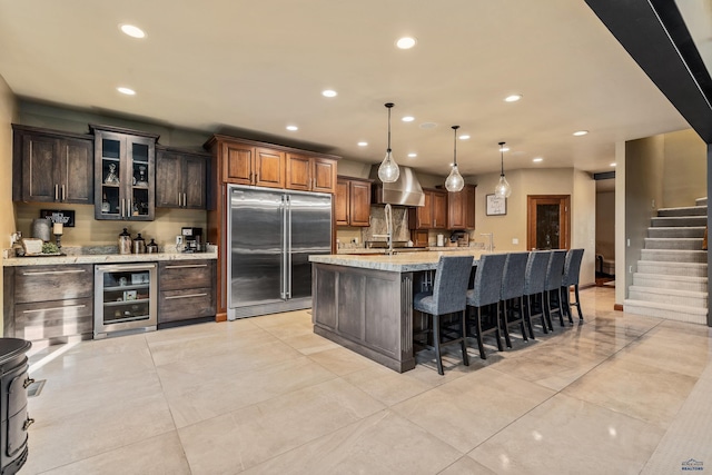 kitchen with hanging light fixtures, beverage cooler, built in fridge, light stone counters, and a center island with sink