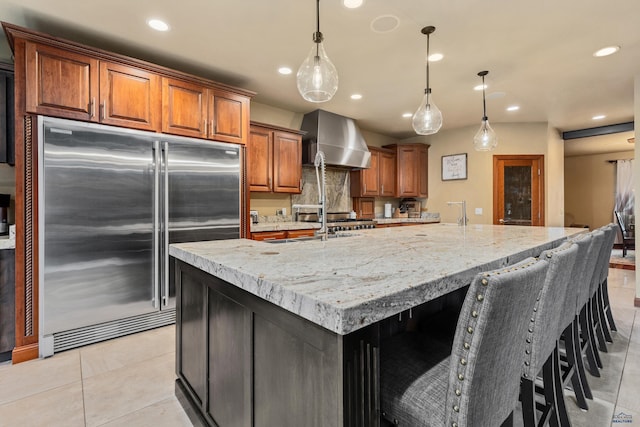 kitchen with stainless steel built in refrigerator, light stone counters, light tile patterned floors, a large island with sink, and wall chimney range hood