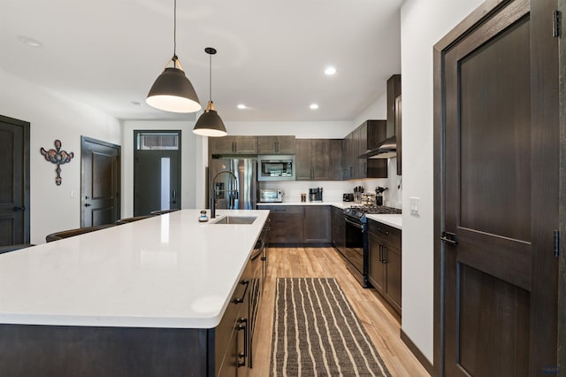 kitchen with dark brown cabinets, light wood-type flooring, an island with sink, appliances with stainless steel finishes, and decorative light fixtures