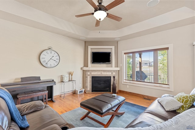 living room featuring ceiling fan, a tray ceiling, a brick fireplace, and light hardwood / wood-style floors