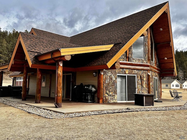 rear view of house featuring stone siding, roof with shingles, and a patio