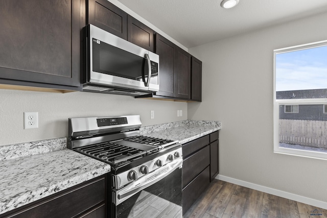 kitchen featuring baseboards, appliances with stainless steel finishes, dark wood-type flooring, light stone countertops, and dark brown cabinets