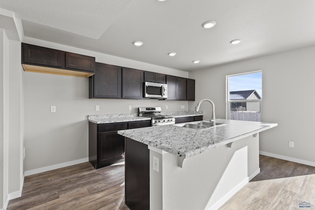 kitchen with stainless steel appliances, a sink, wood finished floors, an island with sink, and baseboards