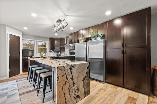 kitchen with stainless steel appliances, an island with sink, a textured ceiling, a kitchen bar, and light wood-type flooring