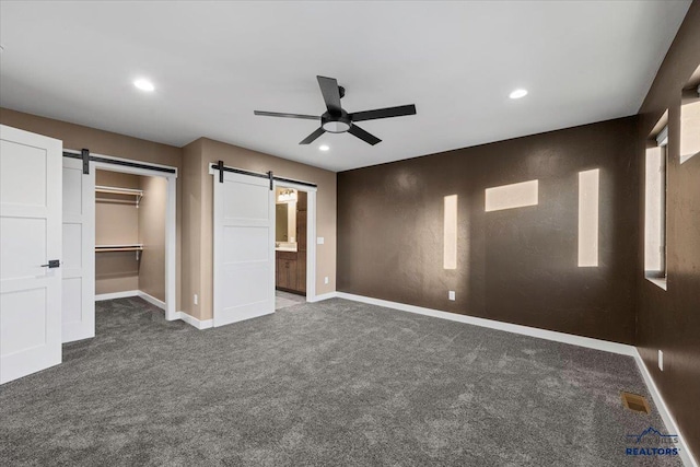 unfurnished bedroom with a barn door, ceiling fan, a closet, and dark carpet