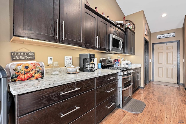 kitchen featuring lofted ceiling, dark brown cabinets, light hardwood / wood-style floors, light stone counters, and stainless steel appliances