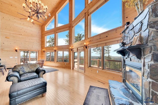 living room with a stone fireplace, high vaulted ceiling, wood walls, ceiling fan with notable chandelier, and light wood-type flooring