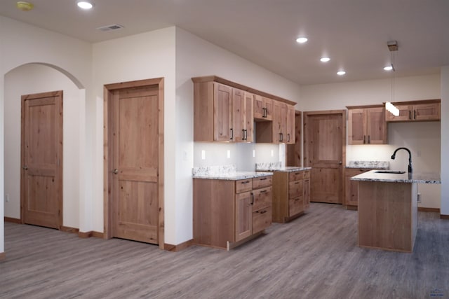 kitchen with light stone countertops, sink, hanging light fixtures, wood-type flooring, and a kitchen island with sink