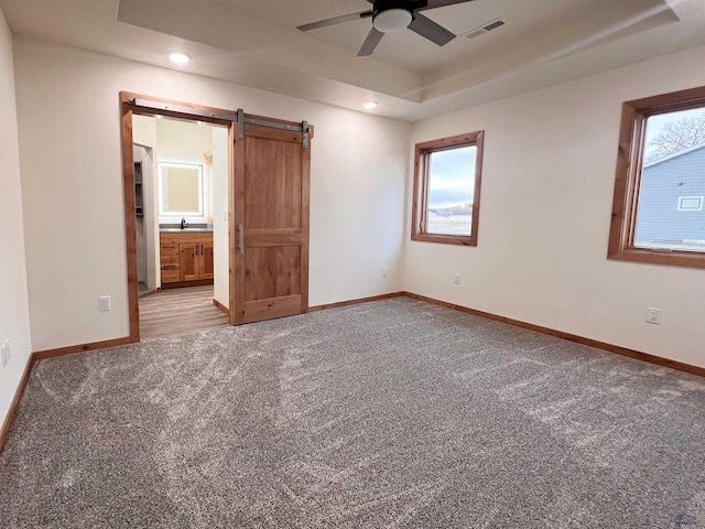 unfurnished bedroom featuring ensuite bathroom, ceiling fan, a barn door, a tray ceiling, and carpet floors