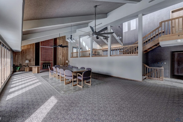 dining area featuring beam ceiling, a brick fireplace, wood walls, carpet floors, and ceiling fan with notable chandelier