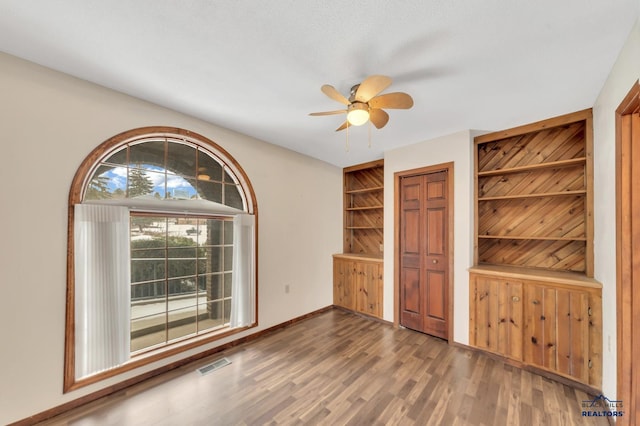 empty room with dark wood-type flooring, built in features, and ceiling fan