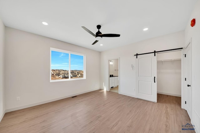 unfurnished bedroom featuring light hardwood / wood-style flooring, a spacious closet, ceiling fan, a barn door, and a closet