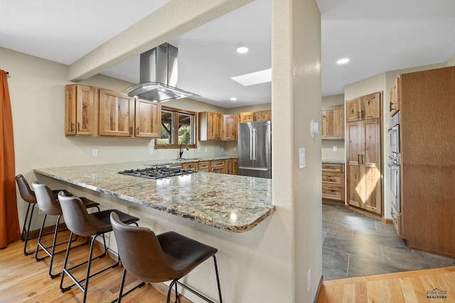 kitchen with appliances with stainless steel finishes, a skylight, light stone countertops, island exhaust hood, and kitchen peninsula