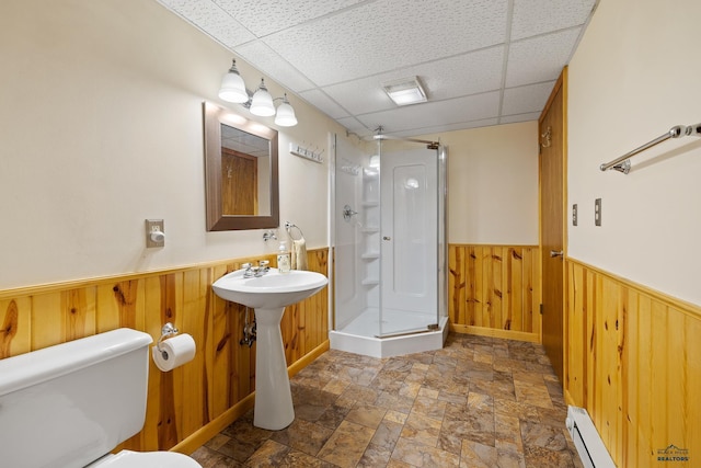 bathroom with a shower with shower door, wood walls, a baseboard heating unit, toilet, and a drop ceiling