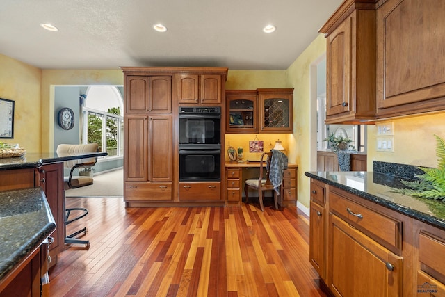 kitchen with double oven, dark stone counters, and light wood-type flooring