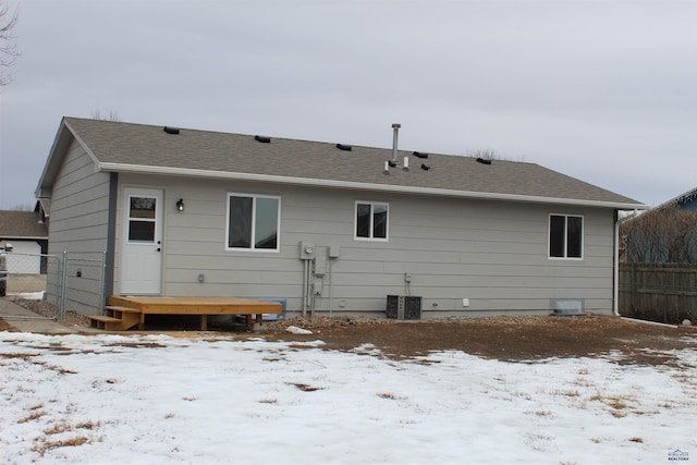 snow covered rear of property with central AC unit, fence, and roof with shingles