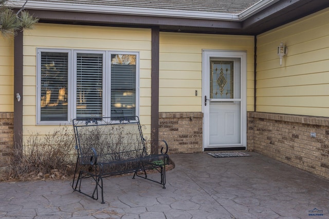 view of exterior entry with a shingled roof, brick siding, and a patio